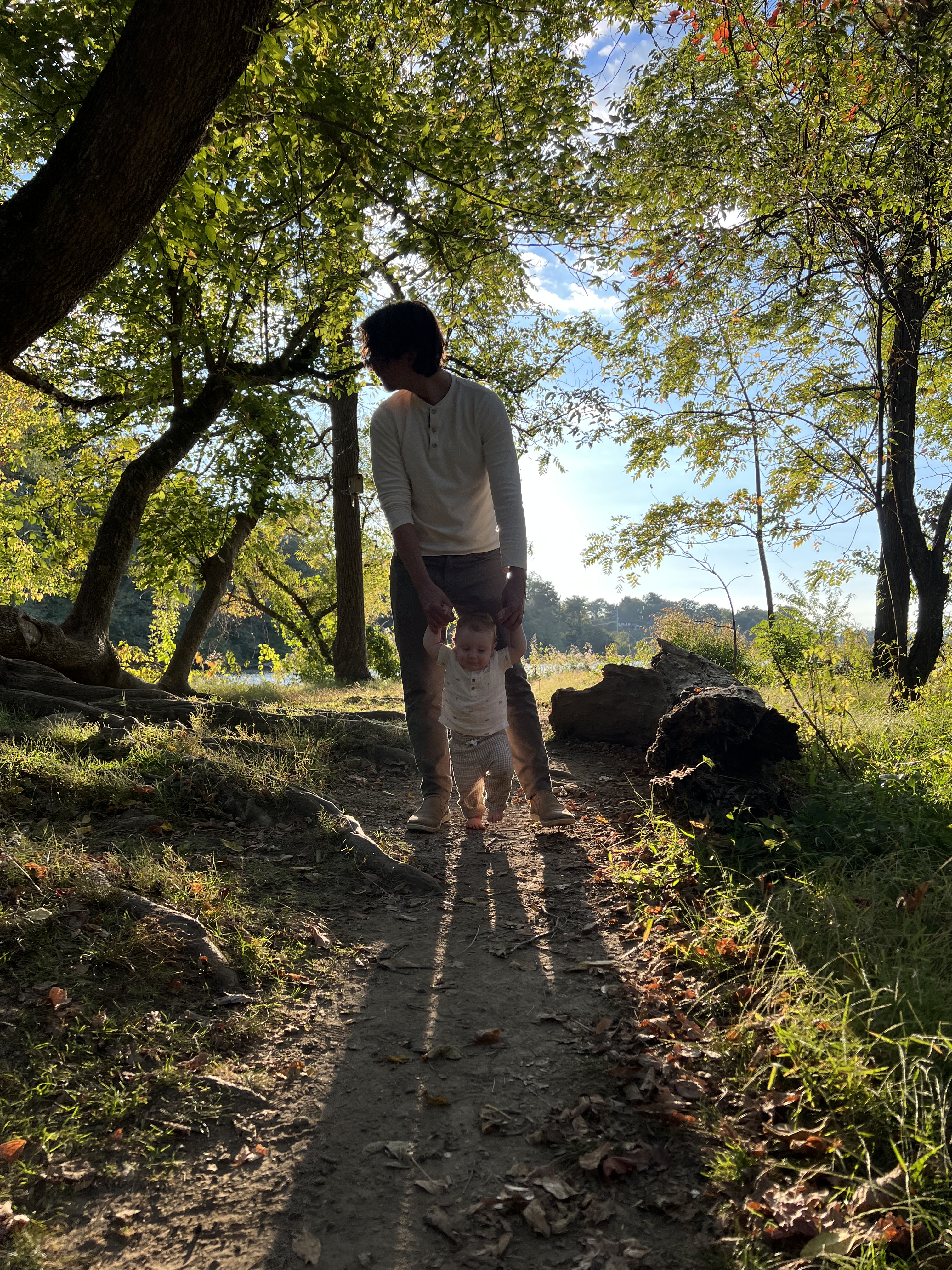 Father helping his young son walk along a wooded path beneath sunlit trees.